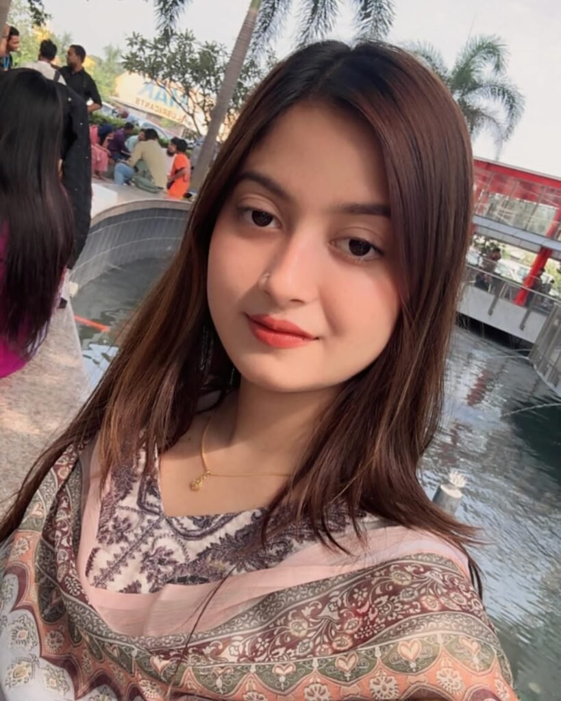 A young woman with long brown hair wearing a patterned scarf takes a selfie by a water fountain, with palm trees and people in the background on a sunny day.
