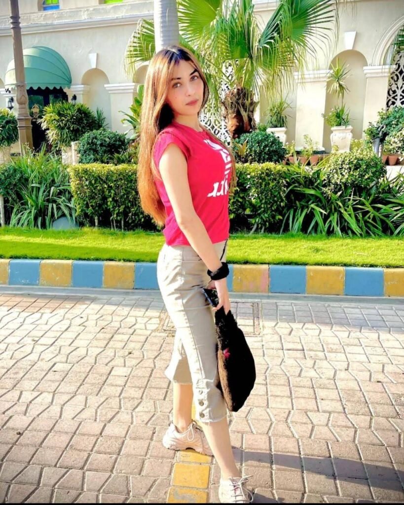 A young woman with long hair, wearing a pink t-shirt, beige pants, and white sneakers, poses outdoors on a sunny day. She holds a black bag, standing on a paved walkway with lush greenery and palm trees behind her.