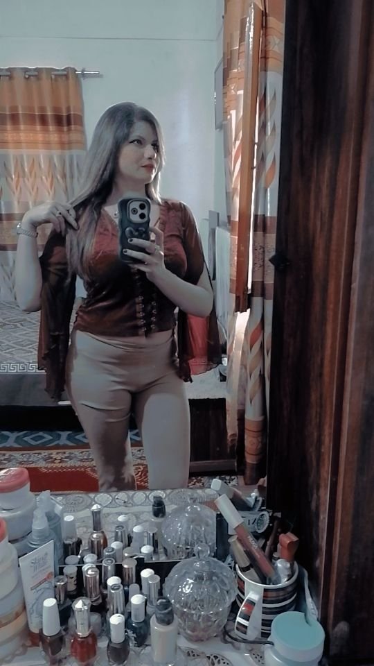 A woman takes a mirror selfie in a bedroom, wearing a brown top and light-colored pants. The vanity in front of her is cluttered with various bottles, makeup, and personal care items