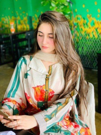 A woman with long brown hair, wearing a floral and striped outfit, sits indoors at a table looking down at her phone. The background features a green wall with yellow flower designs and plants.