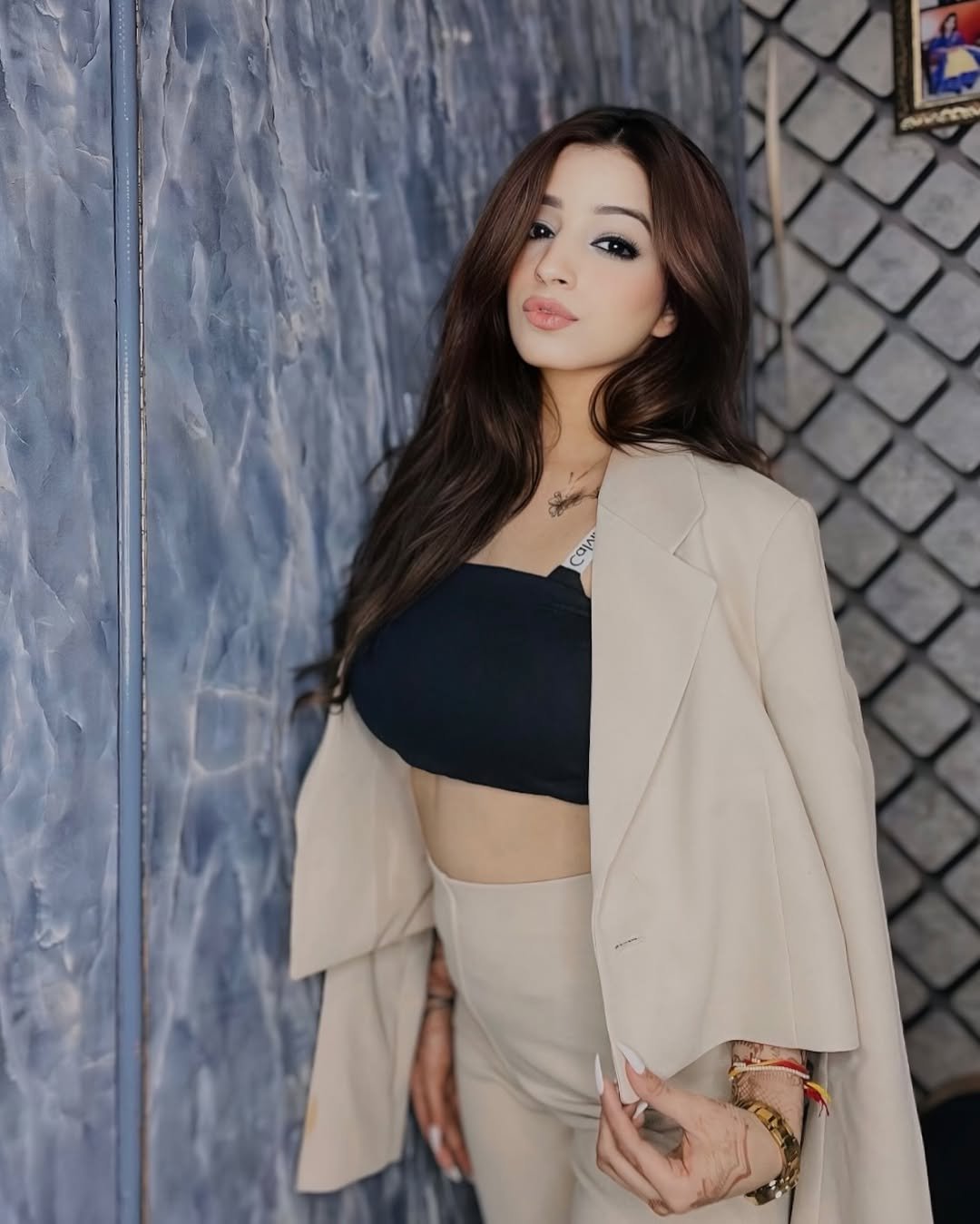 A woman with long brown hair stands indoors wearing a beige suit jacket draped over her shoulders, a black crop top, and matching beige pants. She poses with one hand on her waist against a textured wall.