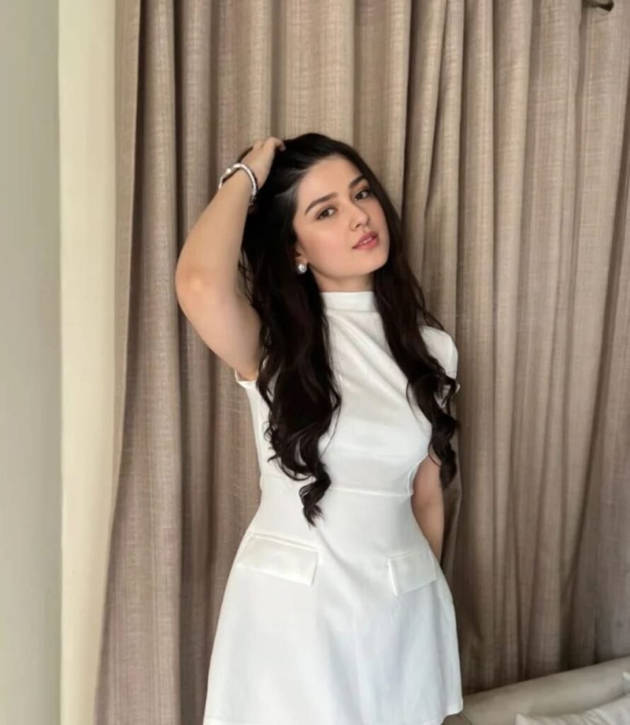 A young woman with long, wavy dark hair wears a short-sleeved white dress and poses indoors with one hand touching her hair, standing in front of beige curtains.