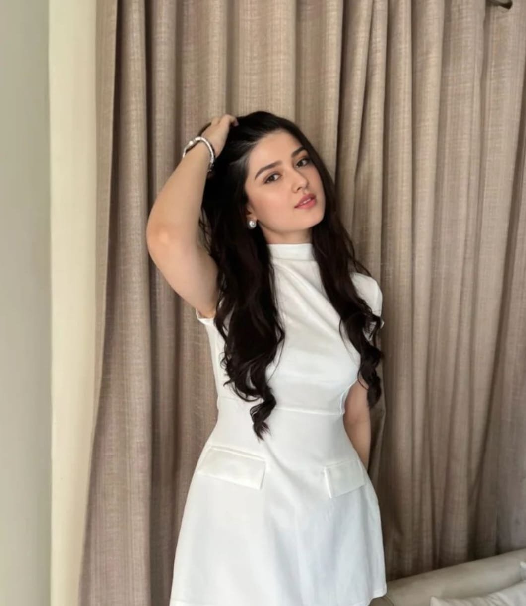 A young woman with long, wavy dark hair wears a short-sleeved white dress and poses indoors with one hand touching her hair, standing in front of beige curtains.