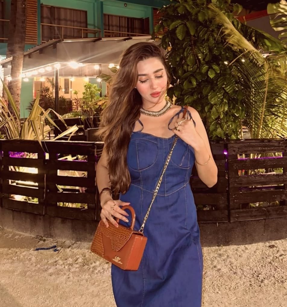 A woman in a blue dress with long hair stands outdoors at night, holding a small brown purse and touching her necklace, with greenery and lights in the background.