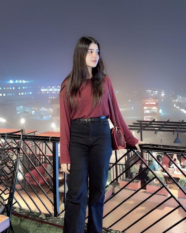 A woman in black pants and a red top stands on a balcony, with a scenic background visible behind her.
